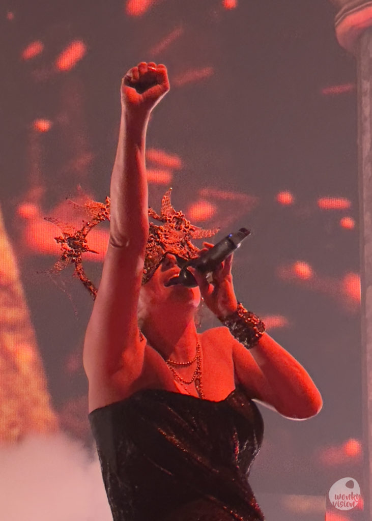 Sharon from Within Temptation sings into the microphone. Her right fist is raised. She wears a crown shaped mask that covers her eyes