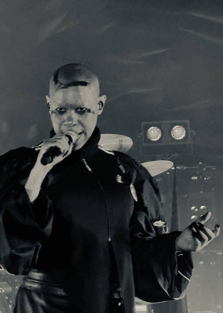 The image shows a Skunk Anansie's singer. She has a shaved head and dramatic makeup. She is singing into a microphone on stage. The lighting is moody, and the photo is in black and white, creating a bold, intense atmosphere.