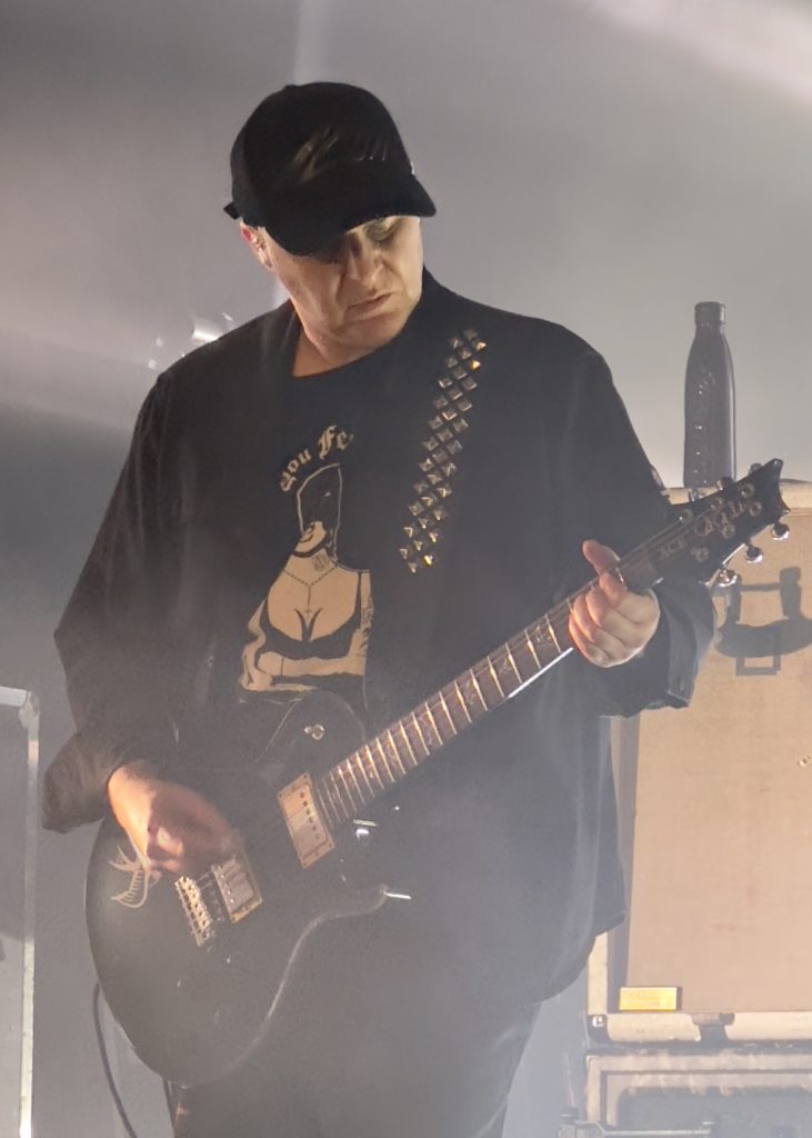 The image shows Skunk Anansie's guitarist on stage, wearing a black cap, black shirt with a bold graphic, and a jacket with metal studs. He is playing a black electric guitar under soft, moody stage lighting.
