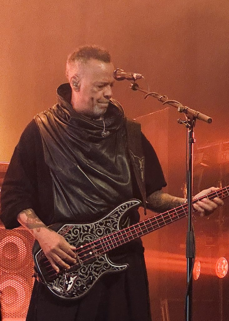 The image shows Skunk Anansie's bassist playing a custom-decorated bass guitar on stage. He is wearing a sleeveless black outfit with a leather hooded top, and has an in-ear monitor. The background is lit with warm orange stage lighting.