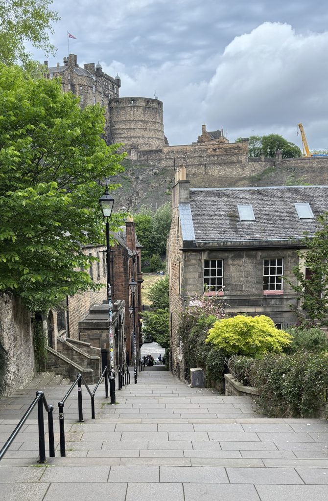 This image shows the famous stepped streets of Edinburgh's Old Town, with traditional Georgian stone buildings lining the descent. Edinburgh Castle dominates the skyline from its dramatic perch on Castle Rock, its medieval towers and walls clearly visible. The scene captures the historic charm of Scotland's capital, with lush greenery and typical Scottish cloudy skies completing the iconic Edinburgh vista