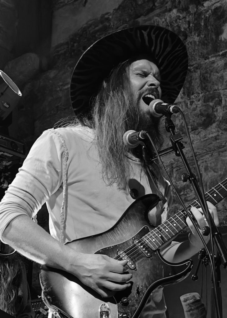 Black and white photo of Jogeir performing live. He has long hair and is wearing a wide-brimmed hat and a light-colored shirt. He is singing passionately into a microphone while playing an electric guitar in an intimate venue with exposed brick or stone walls