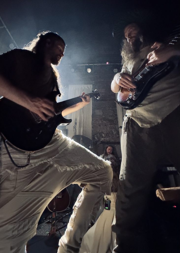 Florian and Jogeir performing on stage in a dramatic, atmospheric setting. The photo is taken from a low angle looking up at two guitarists - one on the left playing what appears to be a bass guitar, and another on the right with long blonde hair playing guitar. The stage is filled with atmospheric smoke or haze, and stage lighting creates a moody ambiance