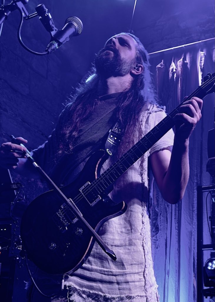 Florian playing guitar with what appears to be a violin bow whilst singing into a microphone above them. The lighting has a dramatic purple/blue tint that creates an atmospheric, moody effect typical of concert photography