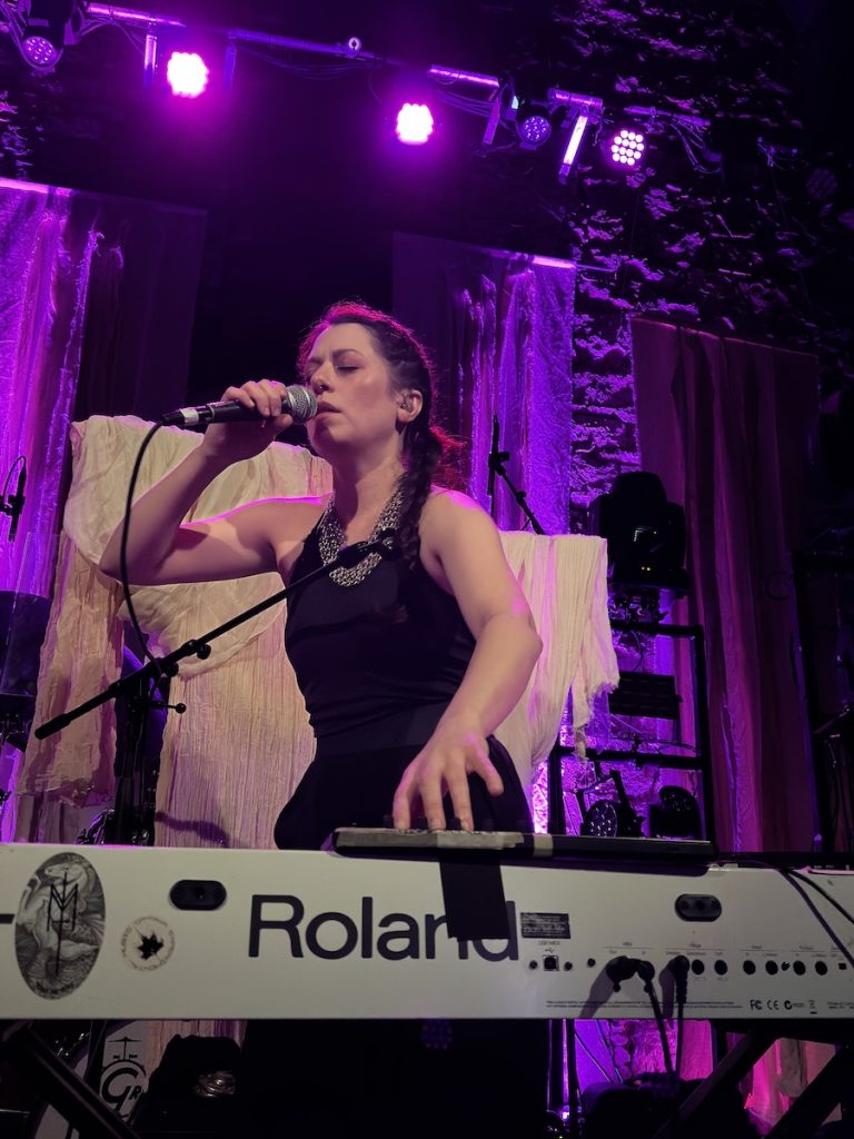 Maud the Moth performing. She's singing into a microphone while playing a white Roland keyboard/synthesizer. She is wearing a black sleeveless top with a decorative chain necklace, and her dark hair is styled in braids.