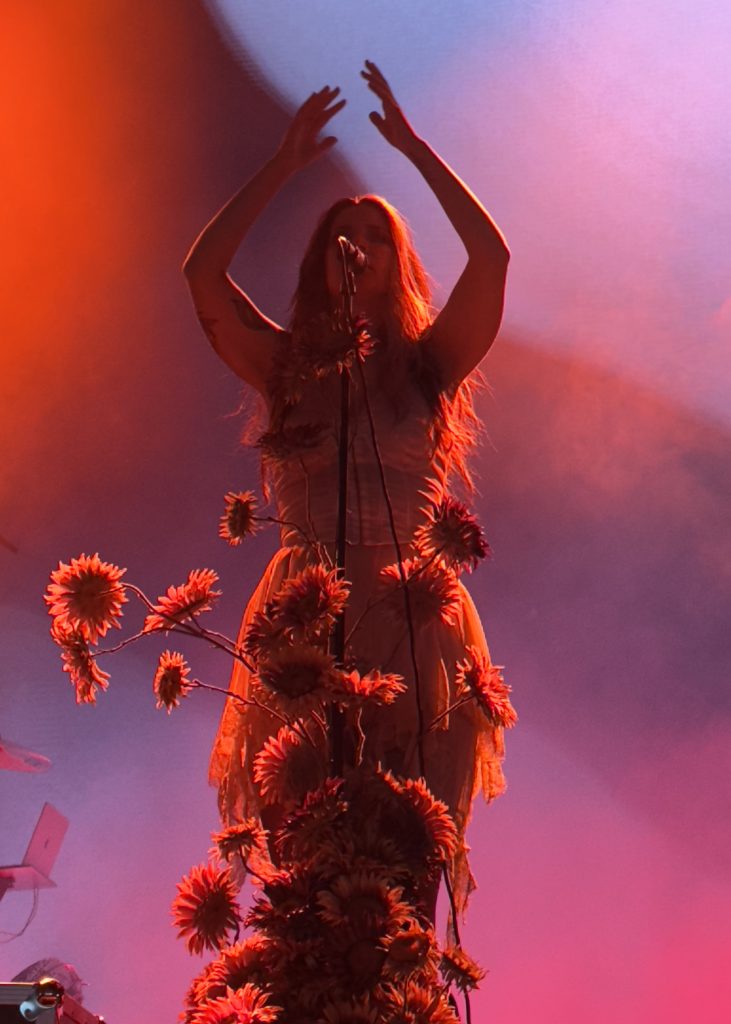 The image shows Charlotte Wessels on stage, singing oon the microphone, with her arms raised gracefully above her head. She is bathed in warm, dramatic reds and orange stage lighting with a haze or fog effect in the background. A decorative arrangement of sunflower‑like blossoms climbs the microphone stand or stage front, adding a vivid, artistic flourish to the performance setting