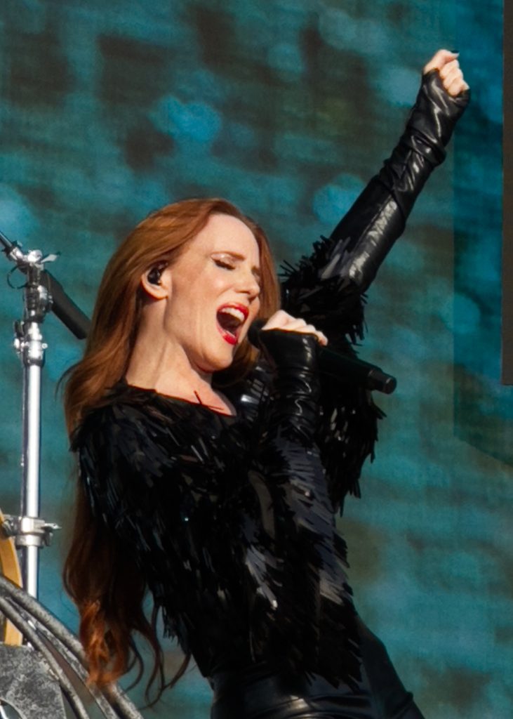 The image shows Epica's lead singer, Simone Simons, holding the microphone and performing passionately, while bold stage lighting and atmospheric effects enhance the visual intensity of the moment