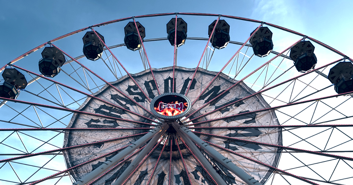The image shows a close-up, upward view of a large Ferris wheel against a clear blue sky. You see the central hub, metal spokes, and enclosed cabins around the top arc. The central part of the wheel has reads Graspop Metal Festival