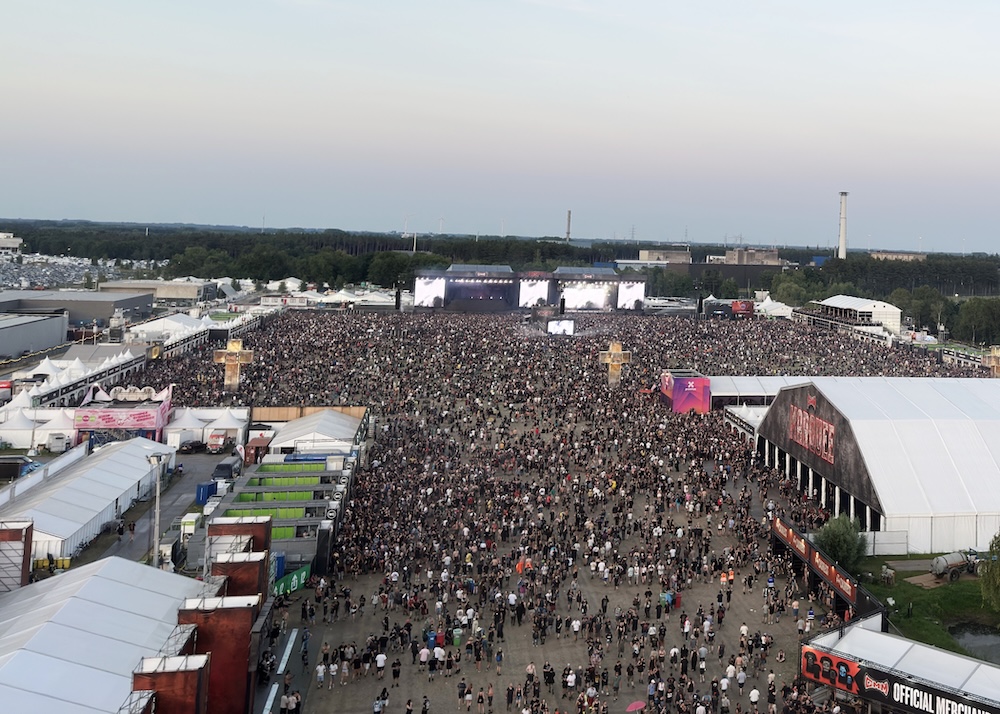 View of Graspop Music Festival from the ferris whell. It shows tens of thousands of people gathered in front of large stages. The crowd fills a vast open area surrounded by tents, vendor stands, and temporary structures, with industrial buildings and forests visible in the background under an overcast sky.