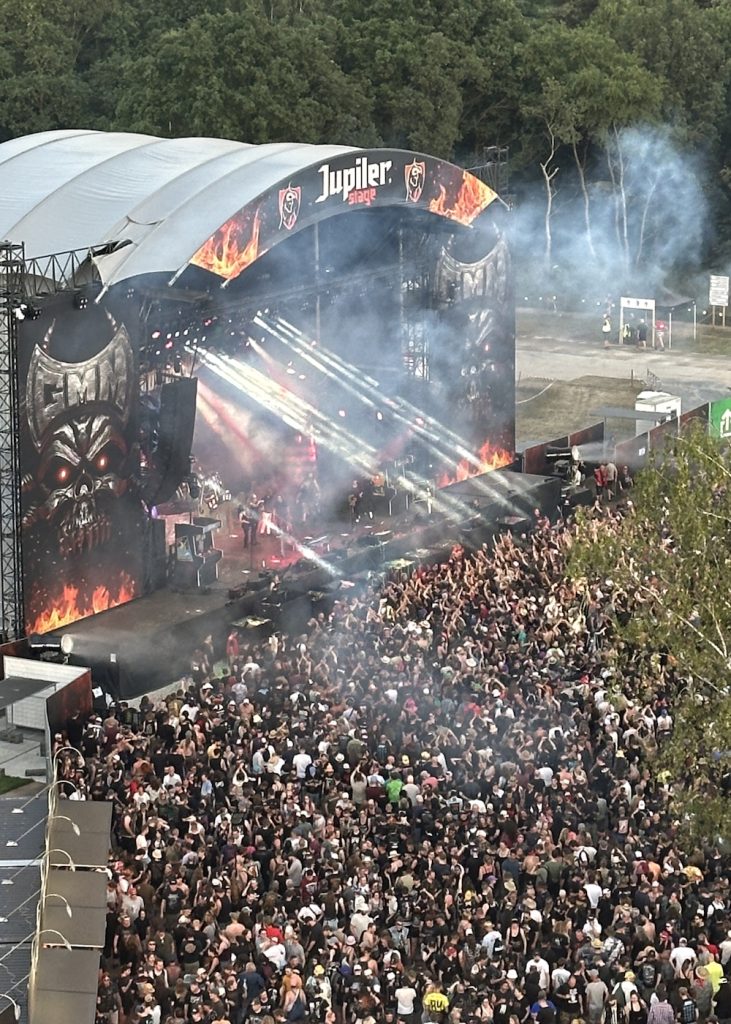 The image shows a large outdoor music festival crowd gathered in front of a stage. The stage has fiery graphics and lighting effects, with a band performing amidst smoke and beams of light. The audience is dense, with many people raising their hands and enjoying the concert.