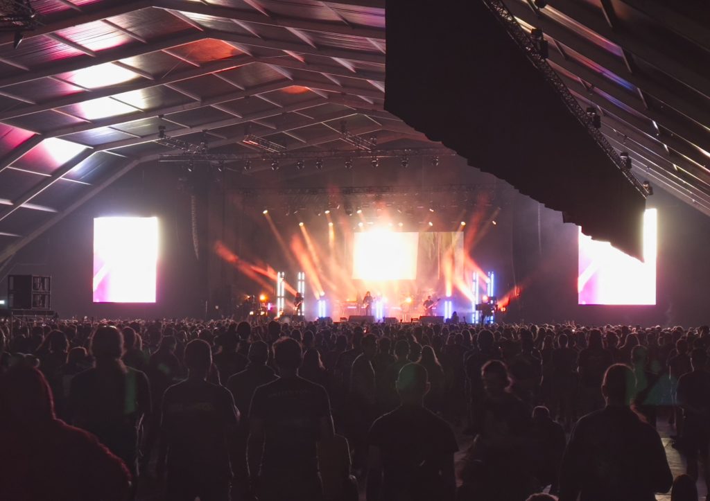 The image shows a live concert scene with Opeth on stage.  Both stage and crowd are on the picture.  There are two screens on each side of the stage.  One of them is partially blocked by short curtains hanging from the ceiling on the satage. The atmosphere is moody and intense, capturing the energy of a metal/rock performance