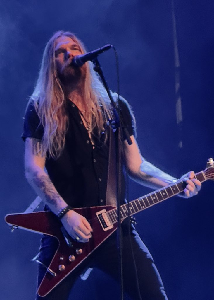 The image shows Lars, on of Soen's guitarist, standing behind a microphone, singing with whilst he plays the guitar. The backdrop is dark and moody with blue and purple tones, and dramatic stage lights cast a spotlight on the performer. A subtle haze adds atmosphere to the moment.