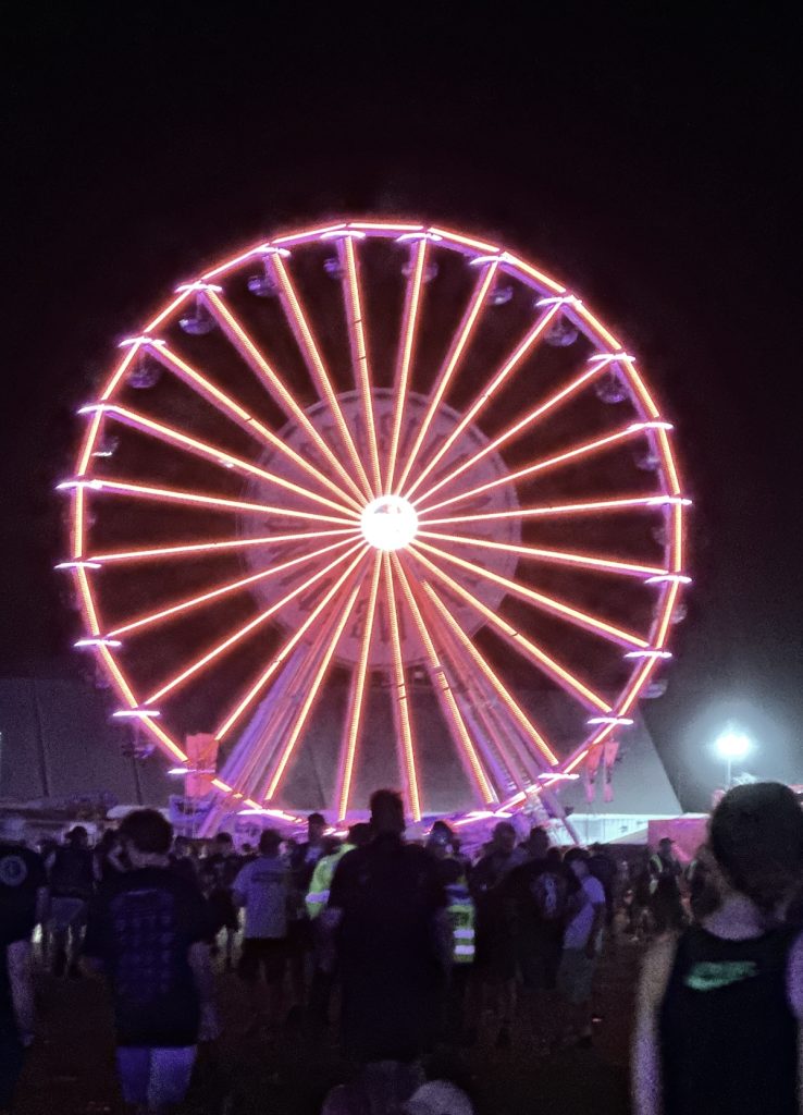 A large Ferris wheel illuminated with pink and purple LED lights against a dark night sky, with silhouettes of people gathered below watching the colorful display