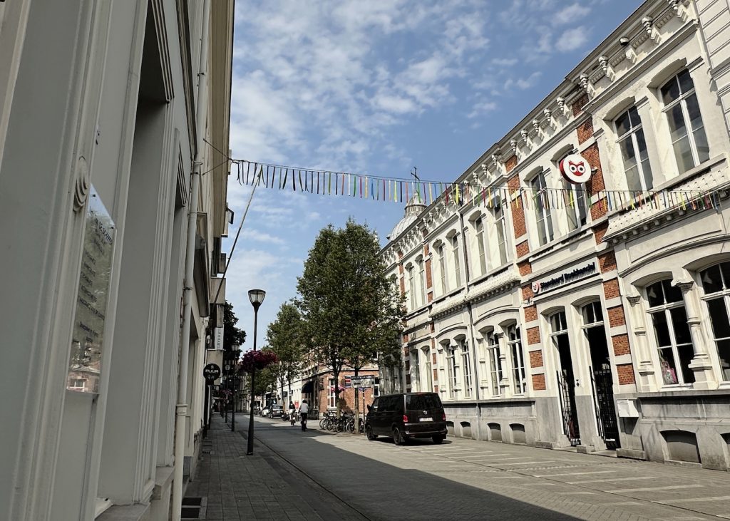 A quiet street of Turnhout lined with elegant historic buildings featuring white and brick facades, decorative bunting overhead, trees, parked bicycles, and a black vehicle under partly cloudy skies
