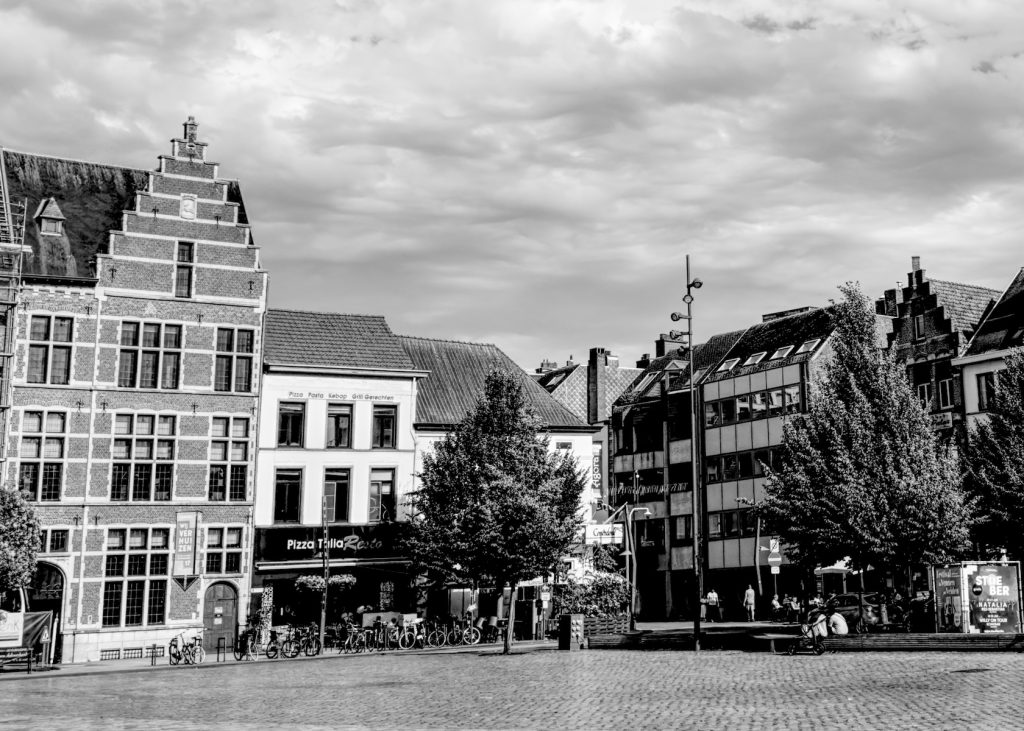 A black and white photo of Turnhout town square featuring traditional stepped-gable architecture mixed with modern buildings, cobblestone pavement, trees, parked bicycles, and a pizza restaurant at street level under dramatic cloudy skies