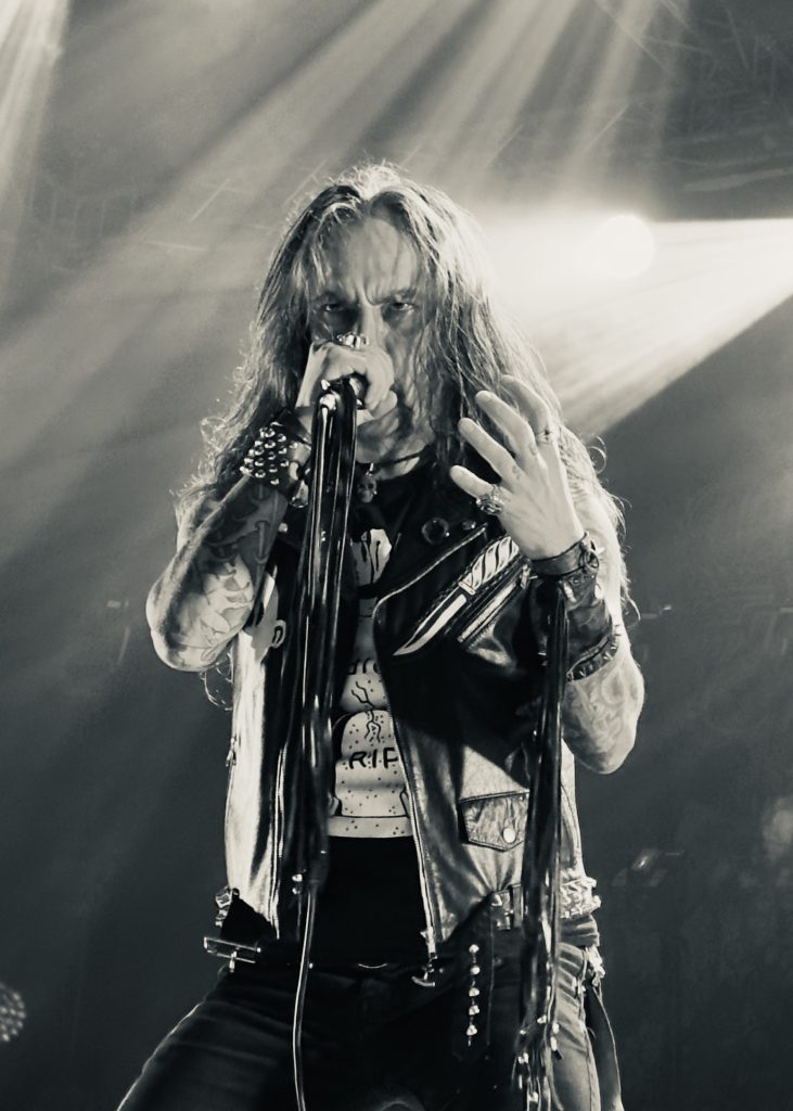 This dramatic black and white concert photograph captures Tomi Joutsen, Amorphis lead vocalist, singing intensely into a microphone on stage. He has long hair and is wearing a leather vest over a graphic t-shirt with studded accessories and bracelets. Shot from a low angle, the image emphasizes the performer's commanding stage presence, with one hand gripping the microphone stand and the other raised expressively. Dynamic stage lighting creates diagonal streaks across the smoky background, adding movement and atmosphere to this powerful performance moment.