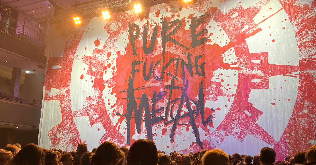 This photo captures a large concert or event venue with a massive stage display featuring bold, aggressive typography. The words "Pure Fucking Metal" are spray-painted in black letters across a dramatic red and white splattered background that resembles paint or blood splatters. The aesthetic is raw with an intentionally chaotic, graffiti-like design.