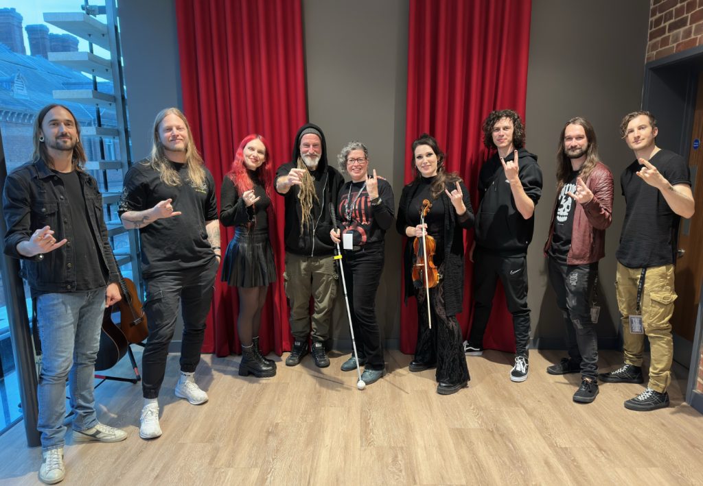 Nivi possing with Eluveitie band members for the VIP photo. She's in the center, holding her white cane. The 8 members of Eluveitie are standing, split in groups of four, on her left and right side. Most of them are making the metal horn sign with their hands.
