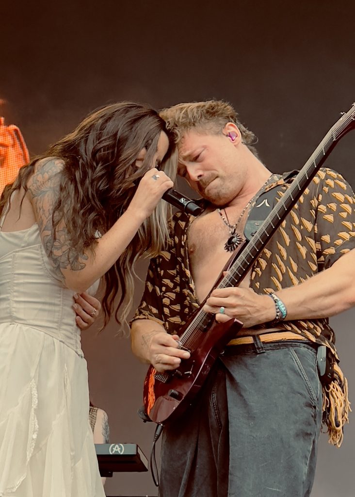 Charlotte Wessels in a white dressleans in close to Timo Sommers who is playing a guitar. She is holding a microphone whils she sings. Timo is wearing an open patterned shirt, they are both deeply engaged in the performance