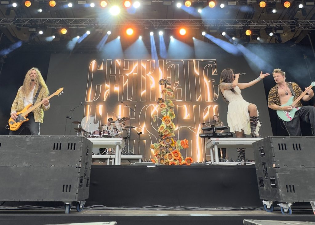 Charlotte Wessels and The Obession performing on a festival stage, with a large illuminated logo backdrop and a mic stand decorated with sunflowers as a centerpiece. Charlotte sings, to her left are Otto (bass) and Timo(guitar), with Joey(drums) and Nina(keyboard / vocals) are visible behind them.