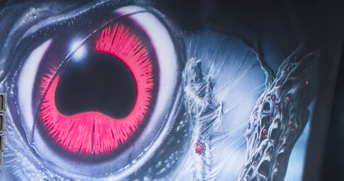 A close-up of the backdrop for Obituary's set. It shows a dark fantasy/horror artwork featuring a large, glowing crimson eye with a black pupil, surrounded by pale, skeletal or creature-like forms against a deep blue-black background