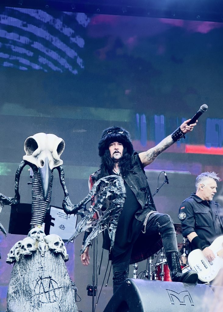 The image shows Al Jourgensen (Ministry's lead vocalists) in dark clothing, fur hat, and spiked wristbands holding a microphone over his head whilst on stage. To his left, there is an elaborate skeletal/skull prop sculpture, Paul Baker (bass) is visible in the background.