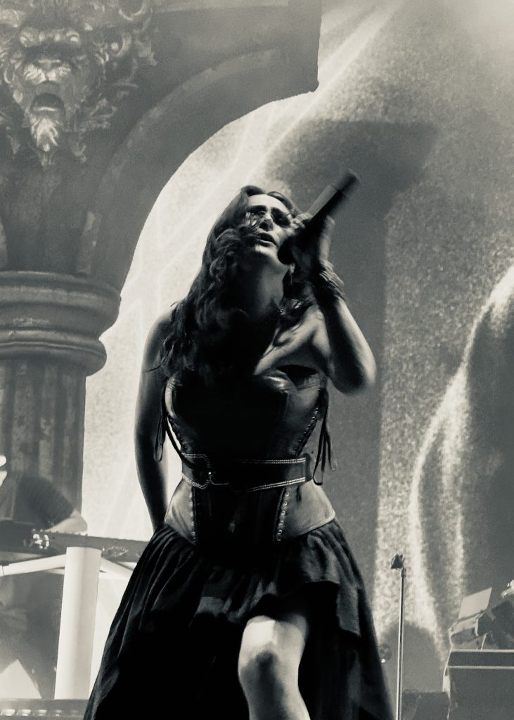 Black and white image of Sharon den Adel (Within Temptation lead vocalist), dressed in a corset and flowing skirt. She tilts her head back singing passionately into a microphone.