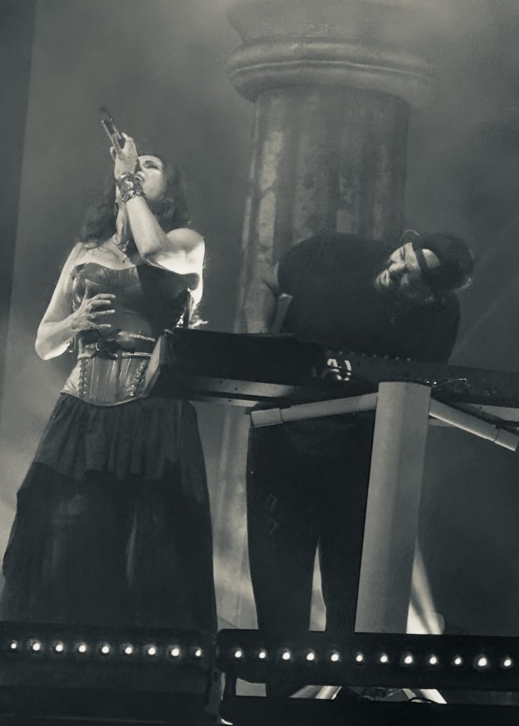 Sharon den Adel of Within Temptation sings passionately into a microphone, while keyboardist Vikram Shankar, wearing a dark cap and t-shirt, leans into his instrument beside her, showing deep emotion whilst performing.